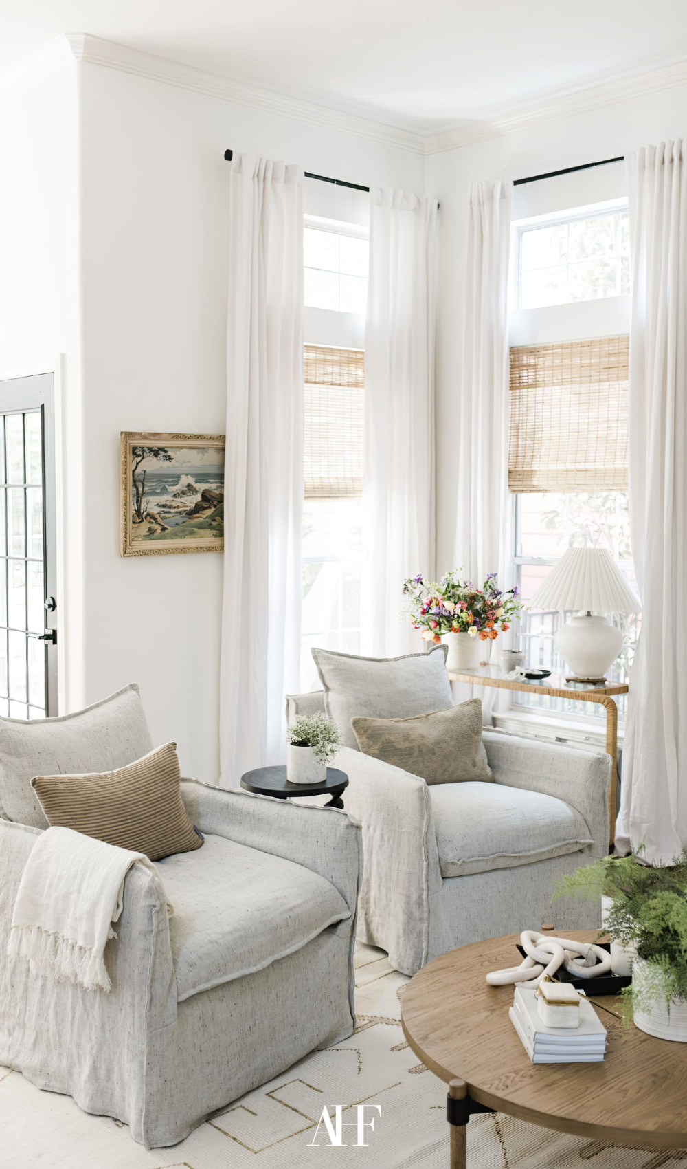 A bright and airy living room with two beige armchairs, white linen curtais and large windows. coastal living room decor.