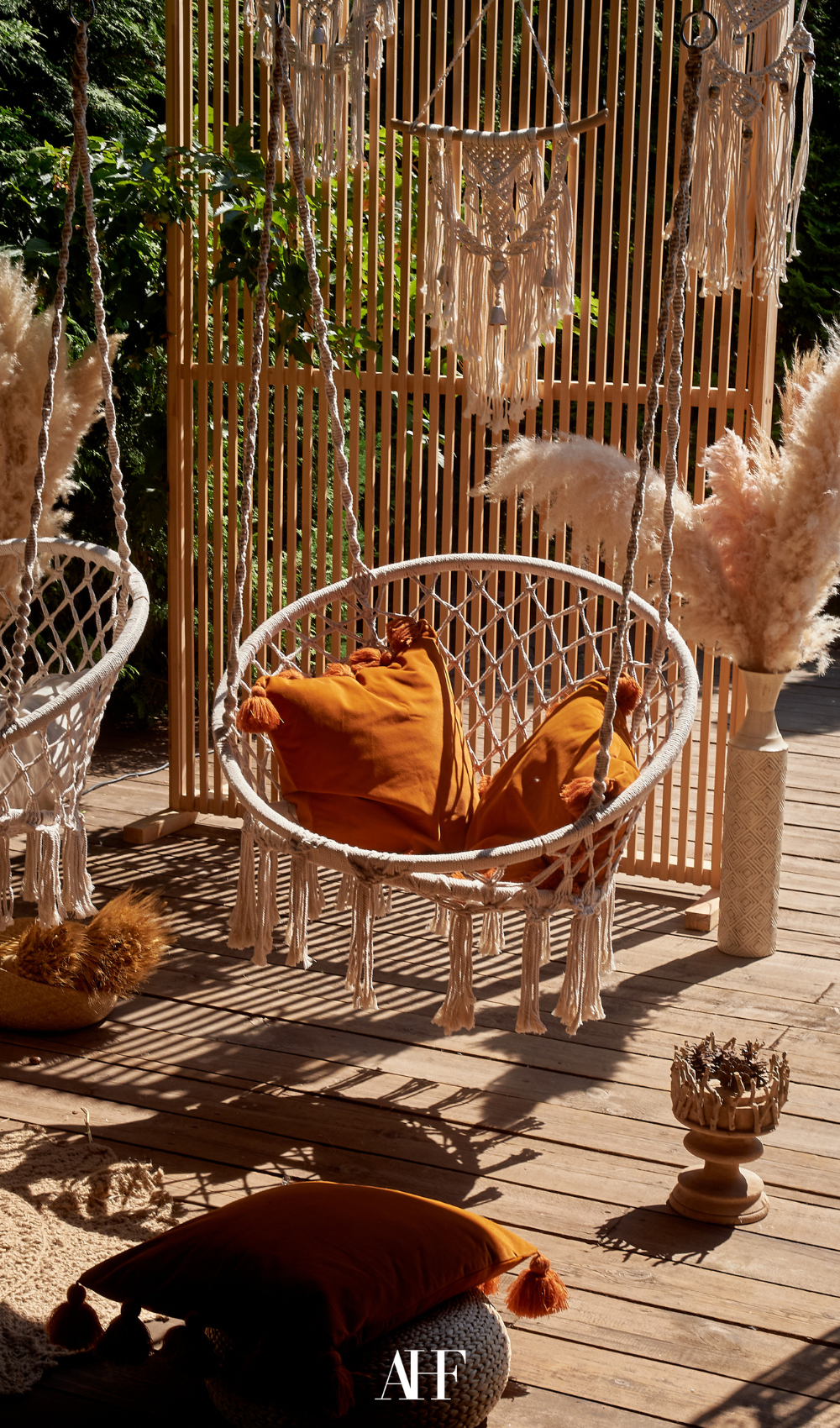 A boho hanging chair with rust pillows inside. Wooden floor and pampas grass in a vase.