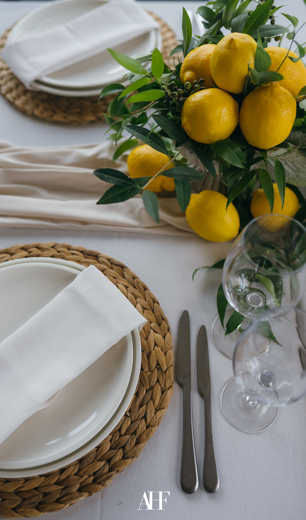 A summer table setting with a white tablecloth, a rattan coaster, white tableware and a bouquet of lemons.