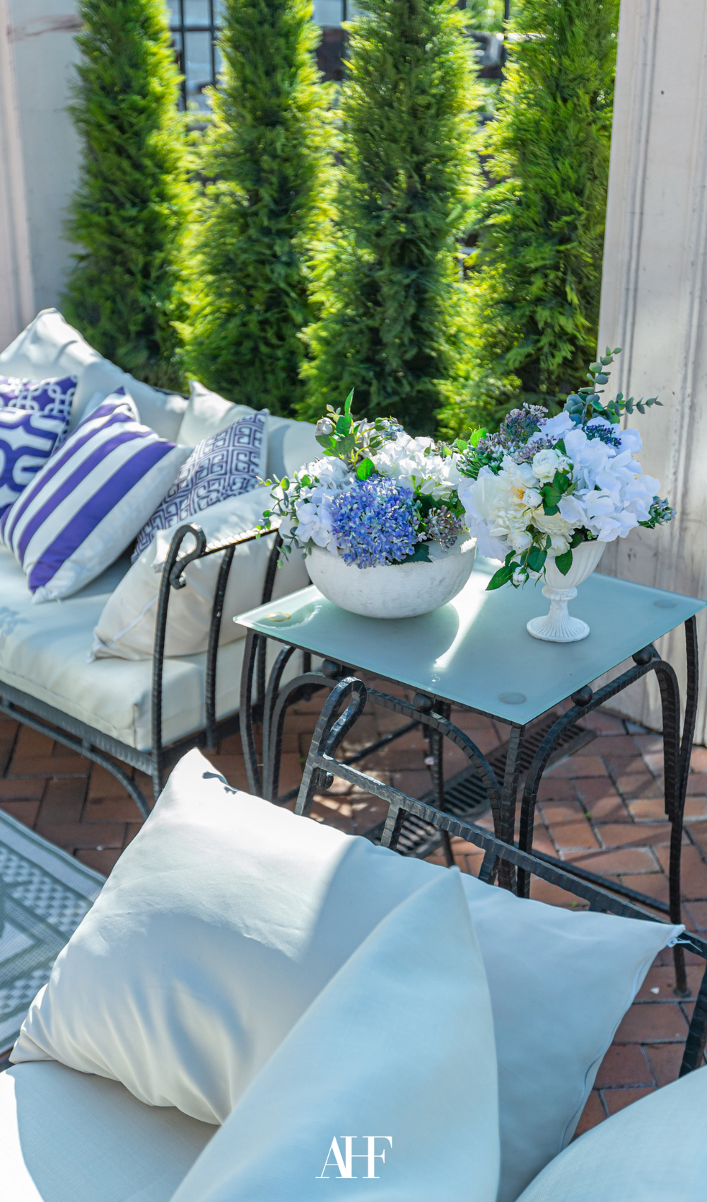 A coastal-inspired small summer patio with white and blue pillows and white and blue flowers. Greenery in the background.