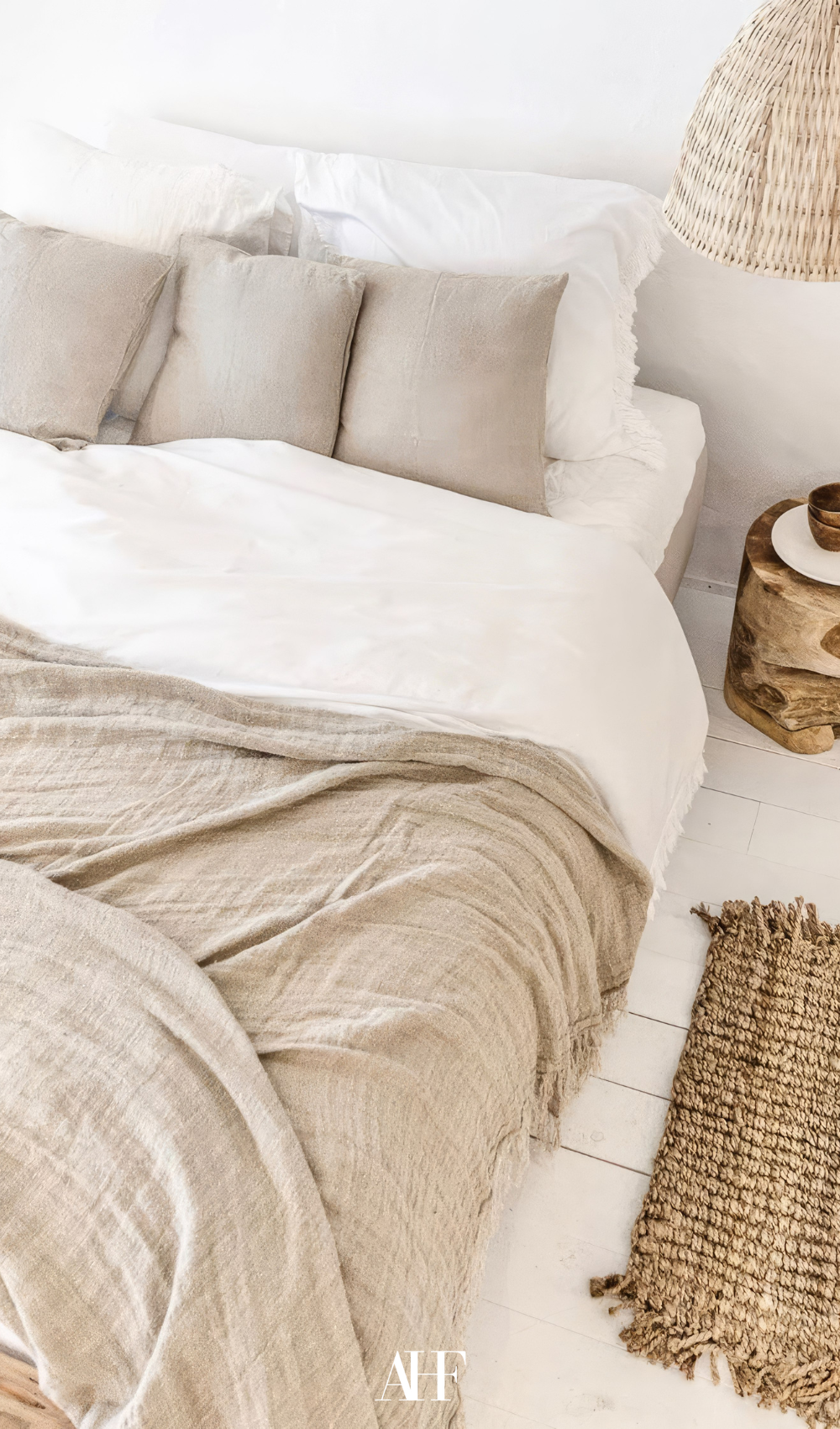 A light bedroom with white bedding and beige blanket and pillows, white wooden floor and a rattan rug.