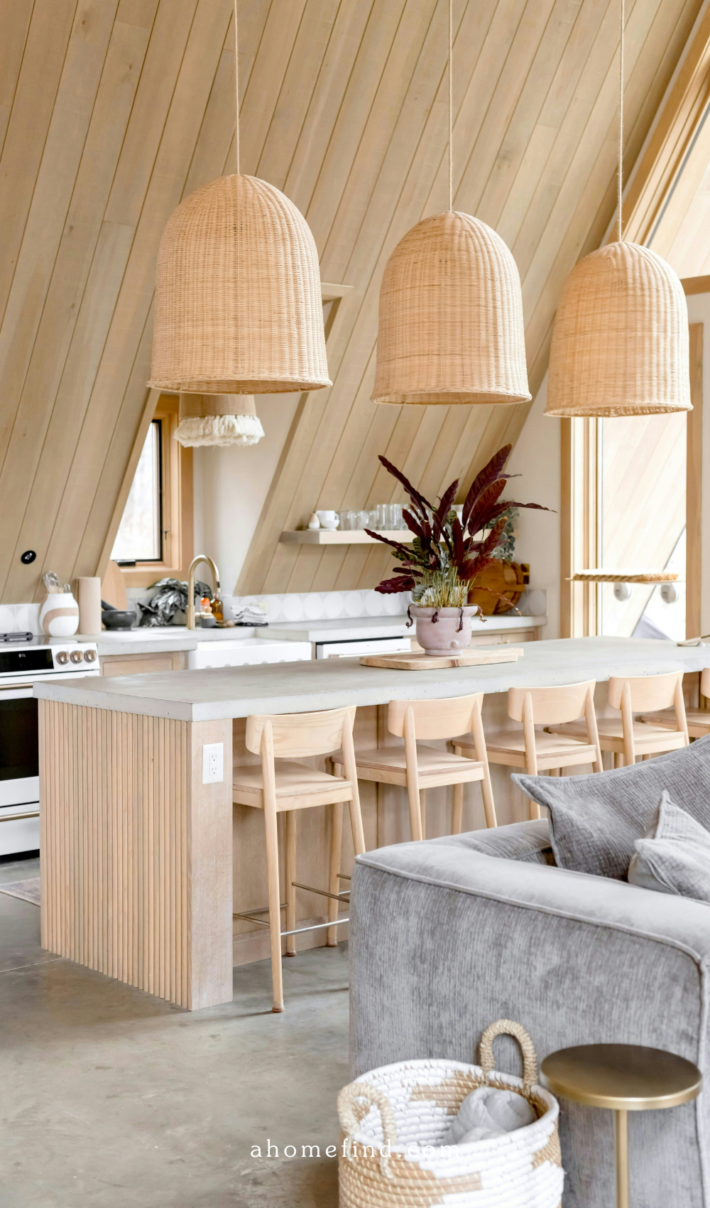 A Scandinavian styled kitchen with light wooden details, three large bamboo lamps above the ktichen island and a grey couch in front of the island. 