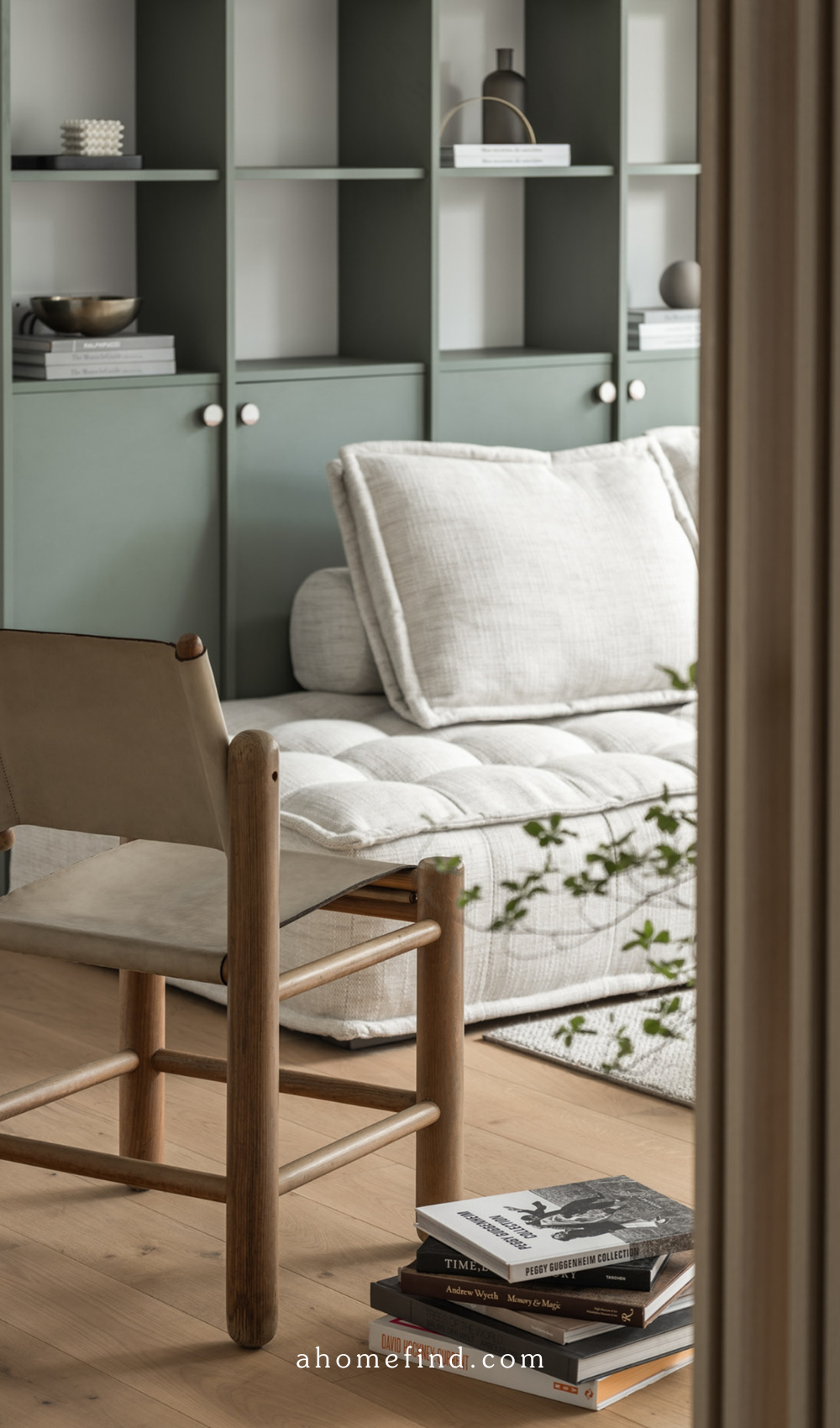 A beautiful white couch with a wooden and rattan armchair in front. In the back is a green bookshelf with storage underneith. 