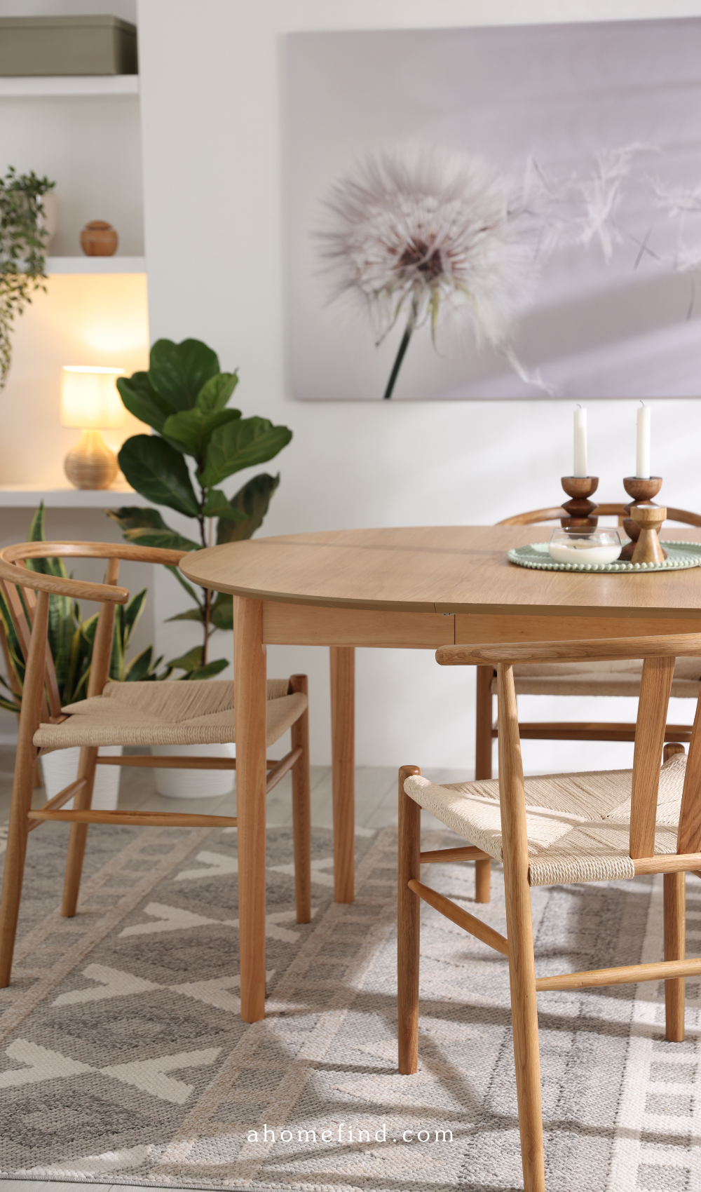 A dining table in a light wooden color, with some decorations. A large plant next to it and a big wall paper. 