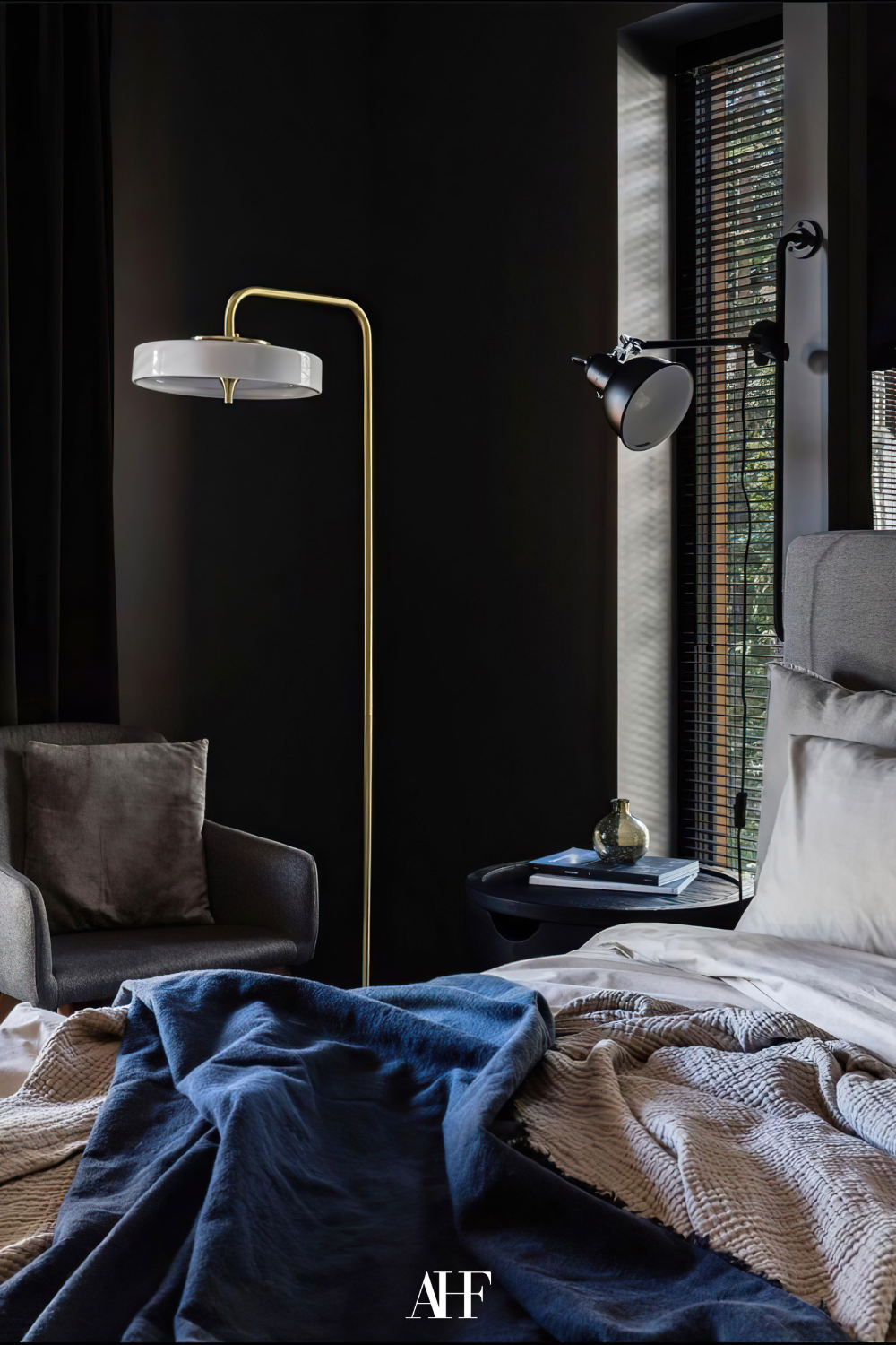 A dark navy and golden bedroom, a window to the right and a bed with a navy blanket. A floor lamp in gold and a darker armchair. 