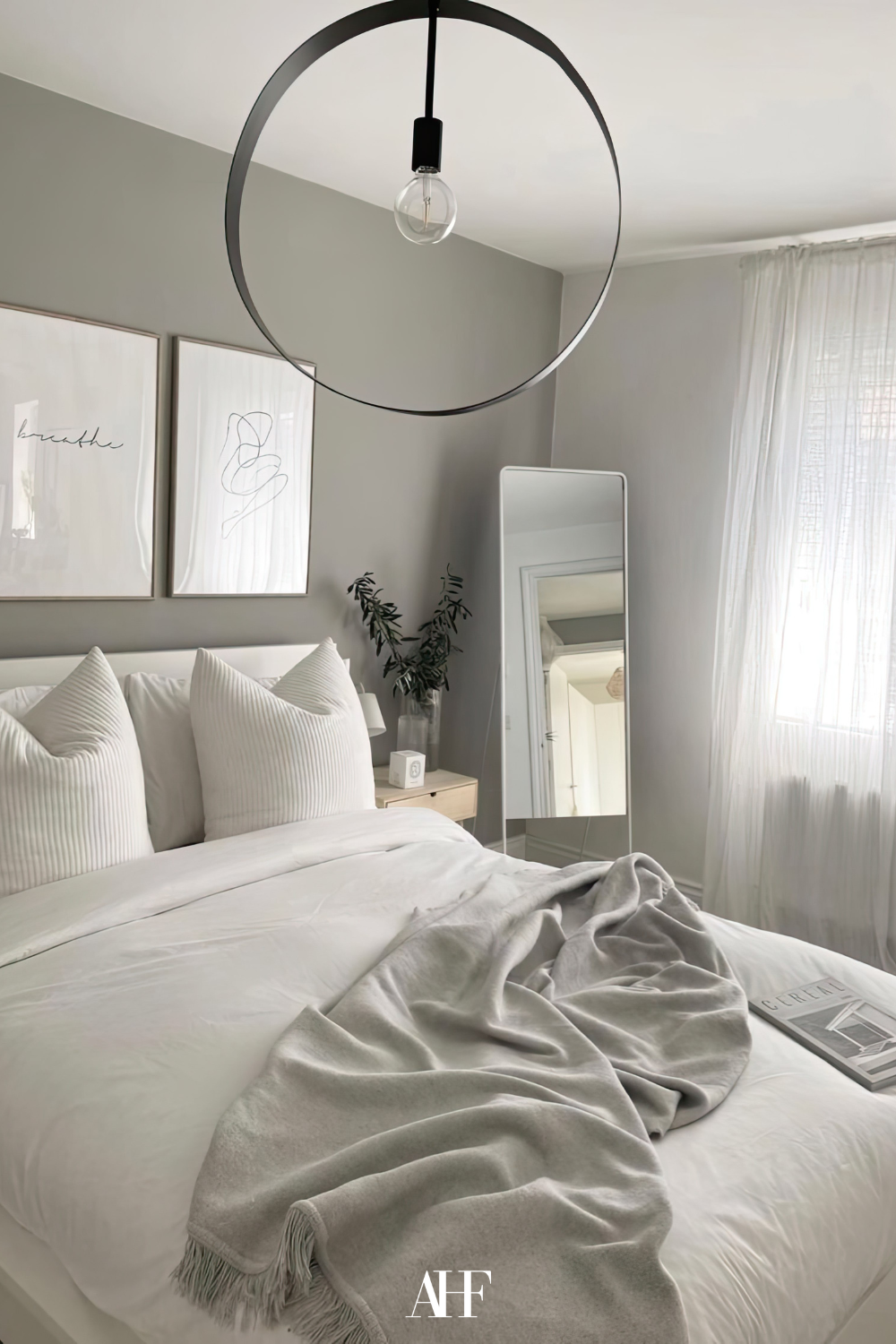 A neutral bedroom color scheme with white bedding, a grey blanket and light grey walls. A large window and a mirror.