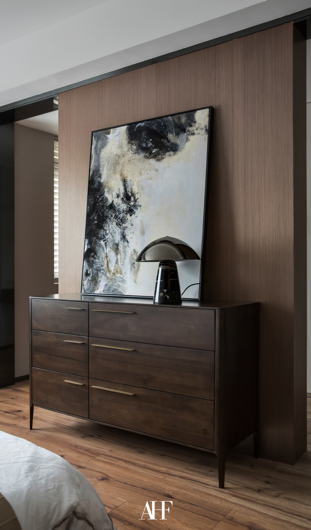 A large black dresser with a black table lamp on it and a large artwork in front of the bed. Dark wooden details.