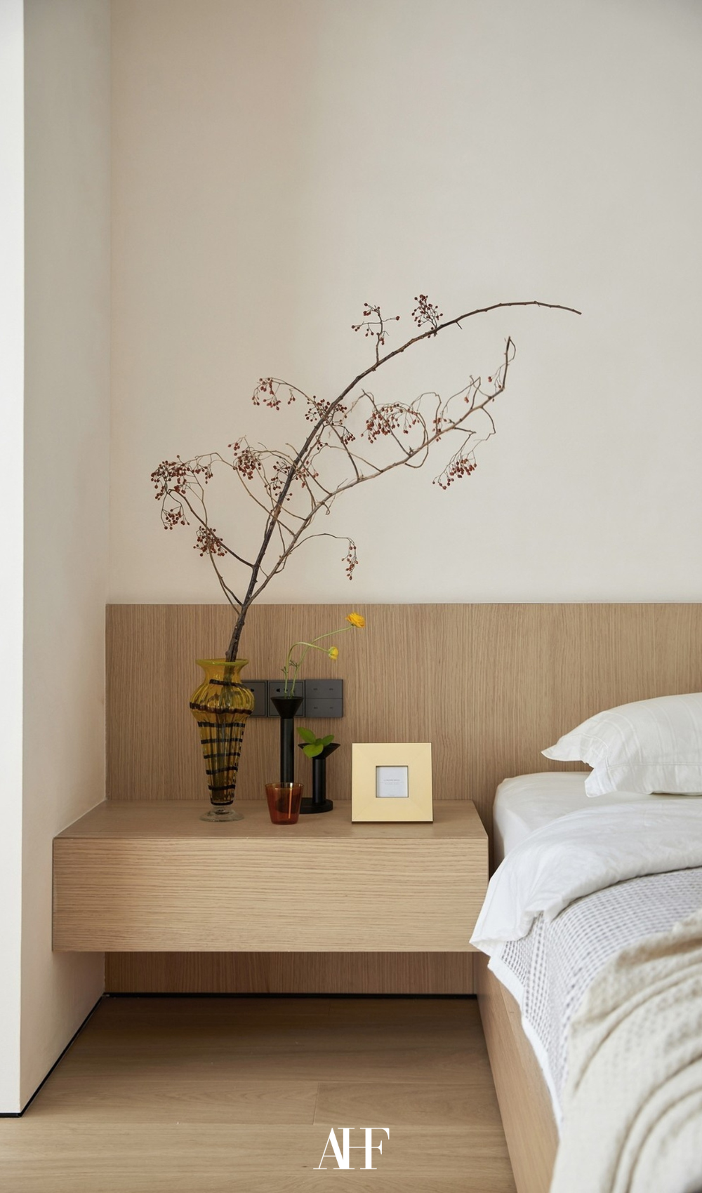 A bedroom with a wooden bed frame and bedside table, light bedcovers and a flower on the bedside table.