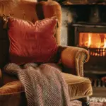 Old money fall vibe with a brown armchair and a rust pillow, along with a An orange sofa with a patterned blanket. A fireplace in the background.