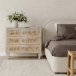 A neutral bedroom with a beige bed and darker bedding, a rattan and white dresser with a plant on it.