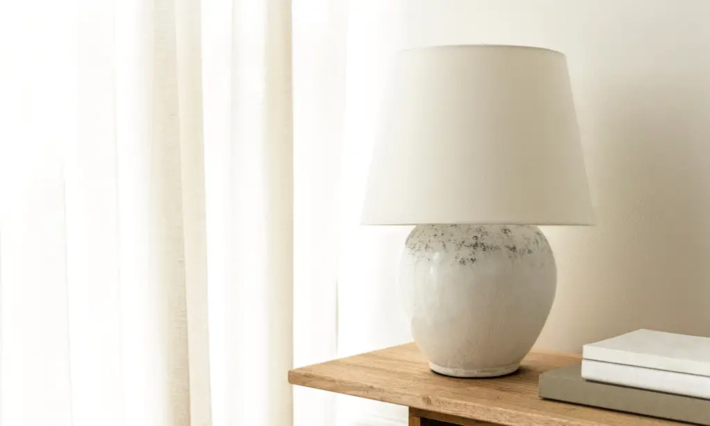 A beautiful large table lamp in a beige color. Standing on a wooden side board with a window to the left.