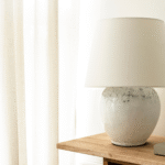 A beautiful large table lamp in a beige color. Standing on a wooden side board with a window to the left.