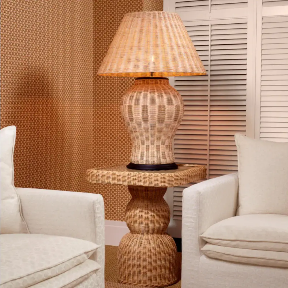 A beautiful rattan table lamp with a black bottom standing on a rattan side table next to two beige linen armchairs. 