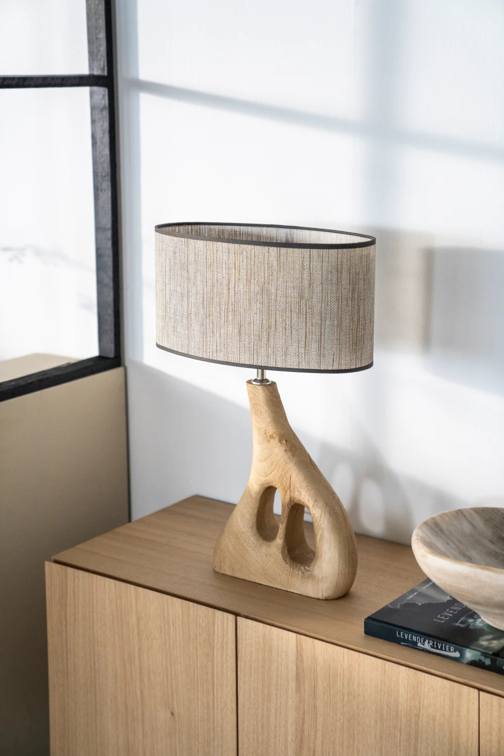 An organic shaped wooden table lamp standing on a wooden side board. A window to the left.