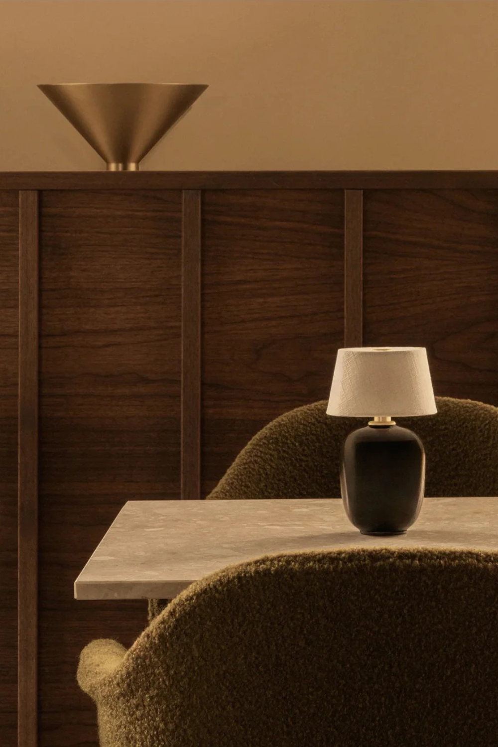 A cozy dark dining room with a dark wooden wall panel, a light marble table and some dark green chairs, along with a dimmable table lamp on the table. 
