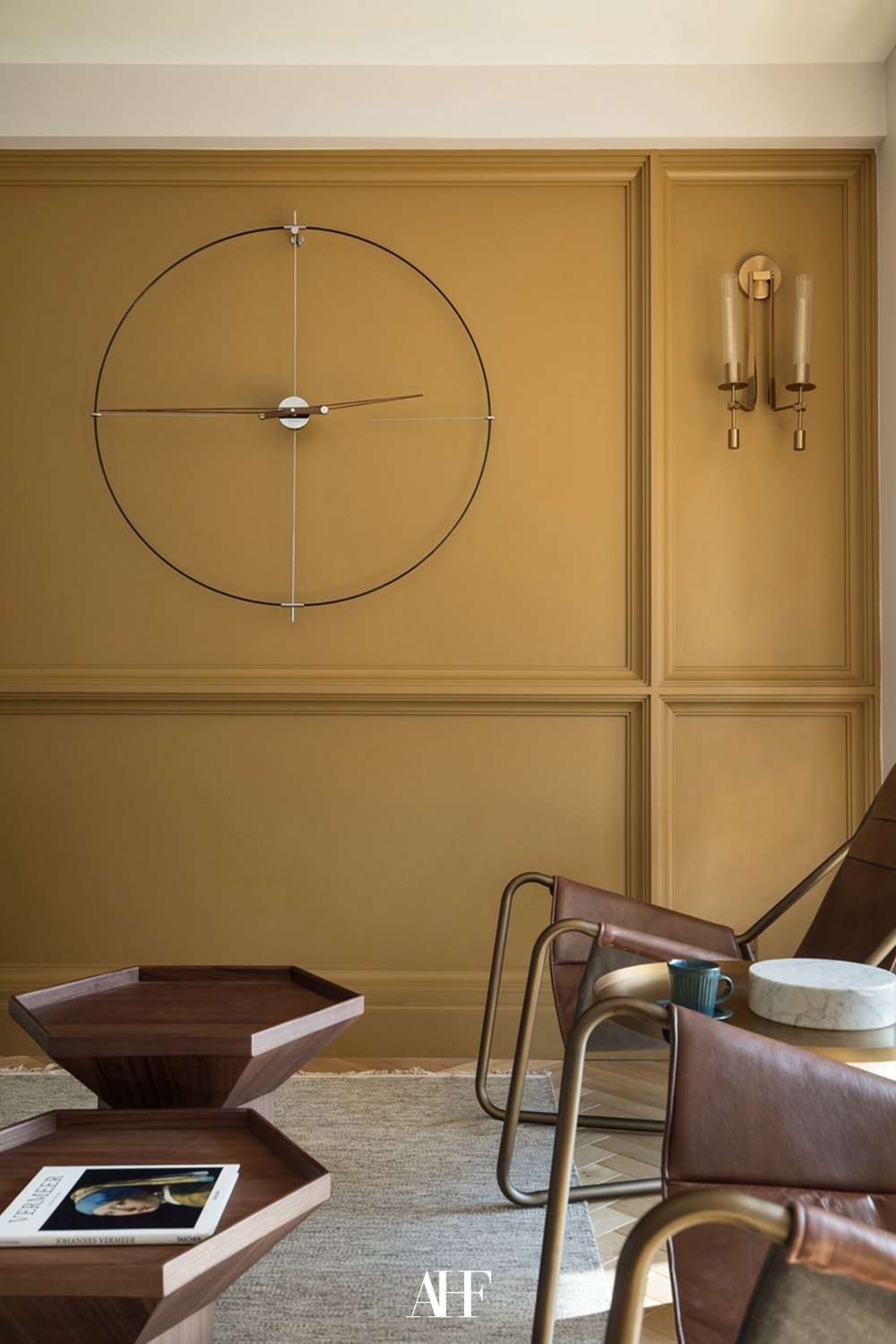 A mustard yellow wall with a large clock and a lamp. A vintage interior design styled room with wooden coffee tables and mid century modern chairs.