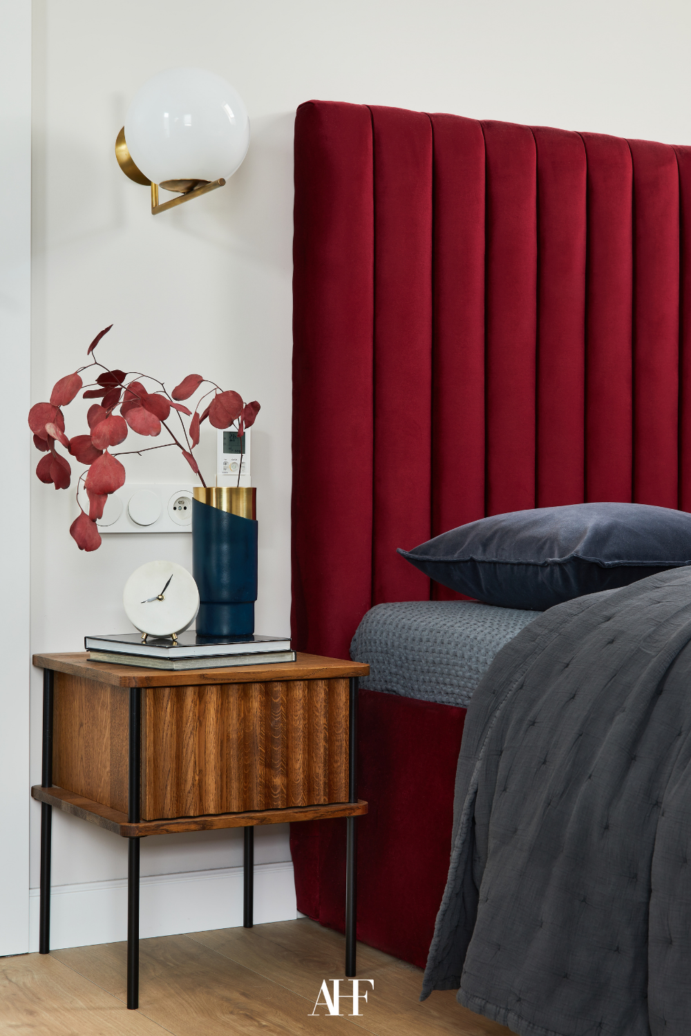 A cherry coded bedroom with a cherry red bed frame and a wooden bedside table. Dark grey bedding. 