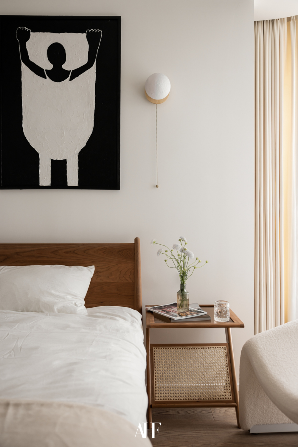 Wooden bed frame with a matching wooden bedside table. White bedding and white curtains. A wall art on the wall.