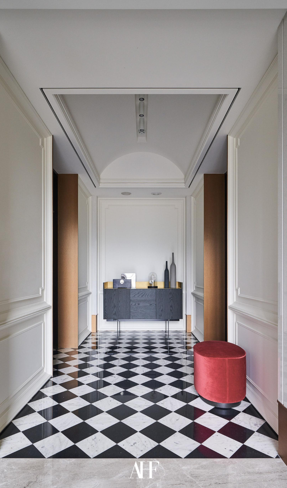 A long hallway with checkered tiles and a console table by the end of the hallway. A red pouf. 